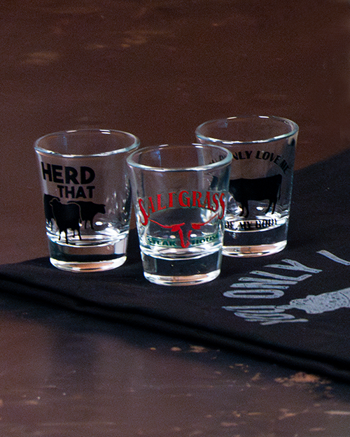 A close image of three shot glasses placed on a dark fabric mat with white letters on a wooden table. Each glass has a unique image of cows, one reading "HERD THAT", another with illegible text, and the one in the center containing the Saltgrass logo.