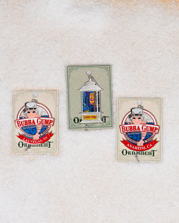 Two round ornaments with the "Bubba Gump Shrimp Co." logo featuring a cartoon shrimp wearing a white hat. The ornament has a red banner at the bottom with 'SANTA MONICA, CA'. Displayed on beige card labeled 'Ornament'. Between the two is another ornament with matching green packaging containing a streetlamp ornament. The background is plain white.