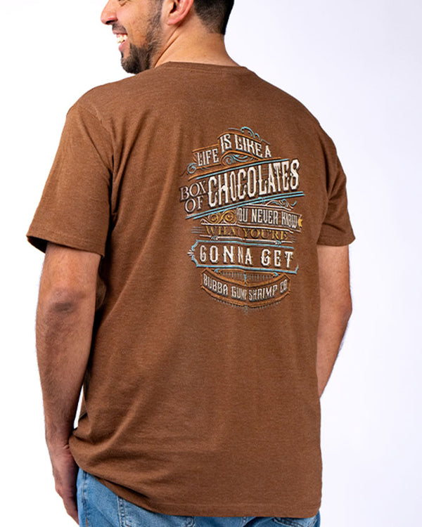 A backside view of a model wearing blue jeans and a brown crewneck T-shirt with a rustic design with linear details, reading 'Life is like a box of chocolates. You never know what you're gonna get. Bubba Gump Shrimp Co.'". The background is plain white.