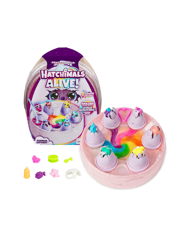 A toy set titled “Hatchimals Alive!” with a Rainbow theme. The set features a circular base with a rainbow-colored swirl at the center and six egg-shaped containers, each showing a small, colorful creature emerging. The packaging displays images of the creatures and includes text such as “AMAZING color change! INCROYABLE! Changement de couleur!” and “RAINBOW HATCHERY.” Several small accessories, including rings, fish, and other miniature items, are also part of the set. The background is plain white.