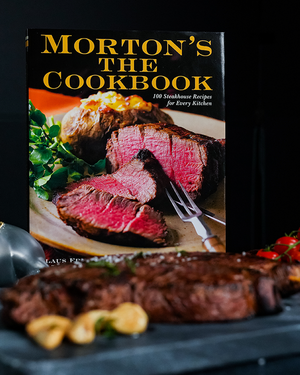 A cookbook titled "Morton's The Cookbook", "100 Steakhouse Recipes for Every Kitchen" by Klaus Fritsch and Tylor Fields with Mary Goodbody. The cover features an image of a sliced steak and a baked potato, with a real cooked steak in front of it on a marble tray. The background is plain white.