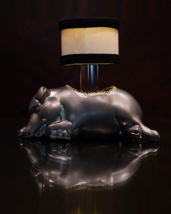 A sideview of a lamp with a silver base shaped like a sleeping pig with a hole in the middle, and a cylindrical lampshade with black trim at the top and bottom coming out of the it. The background is plain black.
