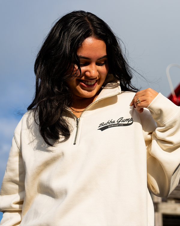 A waist-up view of a dark-haired model against a sky background with her hand up to her chest, wearing a silver ring and a cream-colored 3/4 zip sweatshirt featuring the "Bubba Gump Shrimp Co." logo in navy vintage style cursive text on the left side chest.