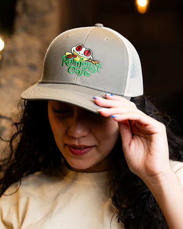 A headshot of a dark-haired model wearing holding with one hand and wearing a white crewneck shirt and a grey and white baseball cap with the text "Rainforest Cafe" embroidered on it and a frog above. The background is plain black and grey.