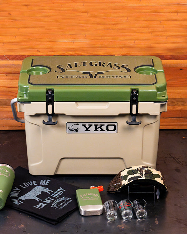 A beige and green cooler with a rectangular silver plate reading "YKO" with a dog head on it, and 'SALTGRASS STEAK HOUSE' and a longhorn head printed on the lid. Surrounding the cooler are a green tumbler, a black cloth with white text reading 'YOU ONLY LOVE ME FOR MY BODY SALTGRASS STEAK HOUSE,' a small green tin, two shot glasses, and a camouflage cap, all placed against a wooden background.