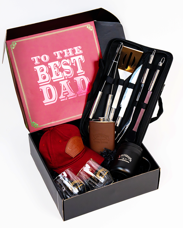 An open black box with various items inside including a red cap, a brown flask, a set of grilling tools in a black case, and two whiskey glasses. The lid of the box has a pink card with gold trim that reads 'TO THE BEST DAD'. The background is plain white.