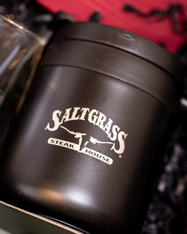 A black tumbler mug with the logo "SALTGRASS STEAK HOUSE" and a longhorn head in the center. The mug is placed in a black box with red and black fabric.
