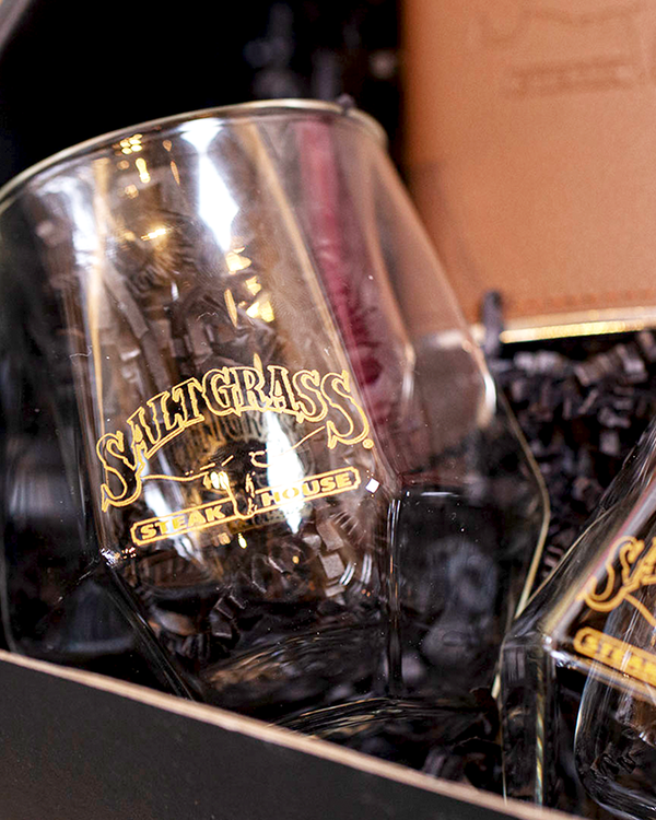 A zoomed in view of an angular whiskey glass with a gold "Saltgrass Steakhouse" logo in the center, next to a matching glass. The cup is placed in black packaging, in front of a brown flask.