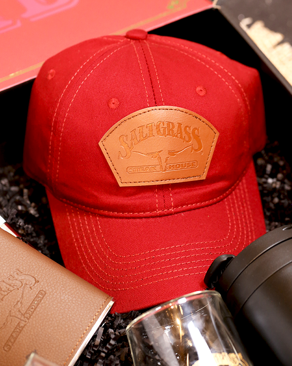 A red baseball cap with a brown leather patch on the front, with the "SALTGRASS STEAK HOUSE" logo embossed on it. The cap is placed in a black box with a flask, a black cup, and a glass.