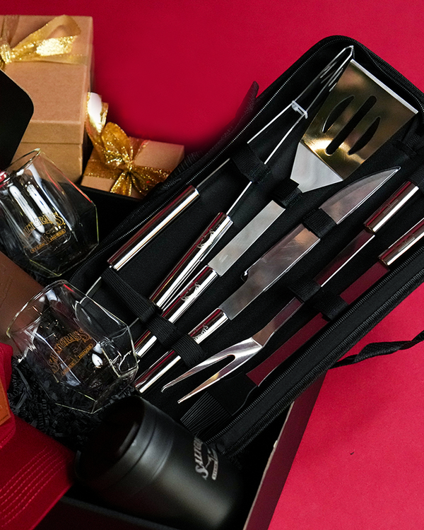 A set of silver barbecue tools neatly arranged in a black carrying case, placed in front of a white background. There are also two angular cut glasses and a black cylindrical container with the word "Saltgrass" in the center. In the background, there are gift boxes wrapped in brown paper and tied with gold ribbons.