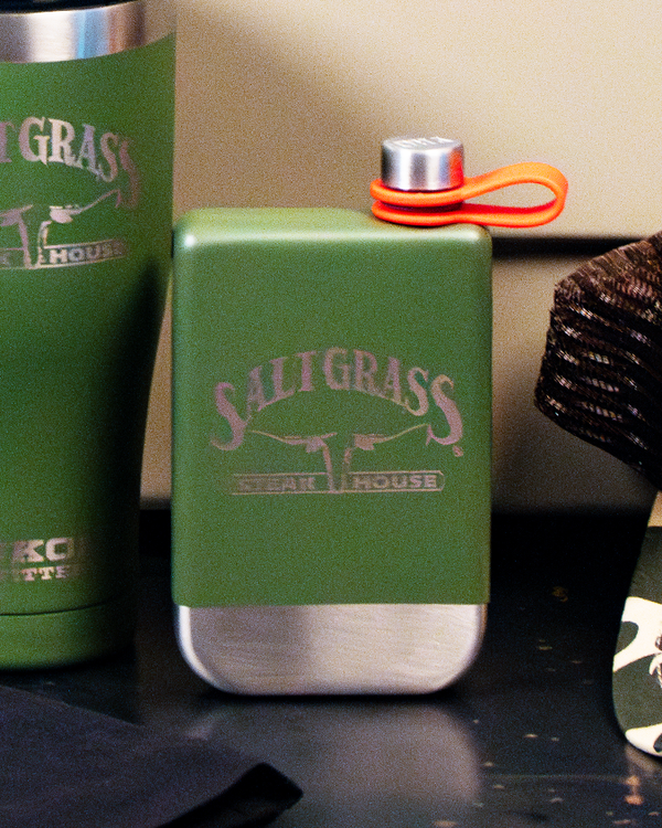 A green flask with a silver bottom and cap with an orange strap attached, and the text "SALTGRASS STEAK HOUSE" printed on the center in silver. Next to the flask is the edge of a matching tumbler cup, and a camo baseball hat on the other side. The background is a black surface and white wall.