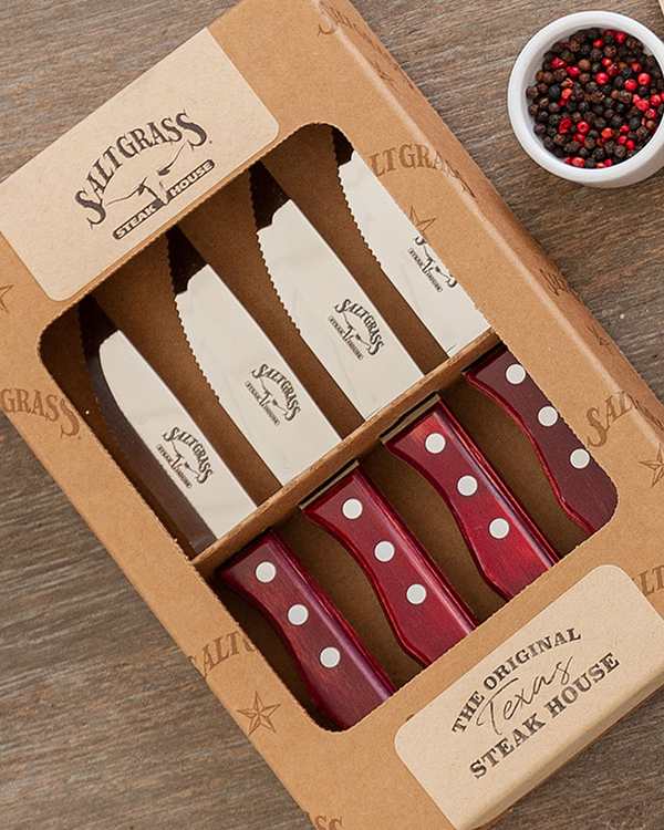 A set of four serrated steak knives with wooden handles, neatly arranged in a box with a cut-out center for visibility, reading 'SALTGRASS STEAKHOUSE', 'THE ORIGINAL TEXAS STEAKHOUSE' at the top and bottom. Each knife blade is engraved with 'SALTGRASS STEAKHOUSE'. The background is a wooden surface with a bowl of peppercorns.