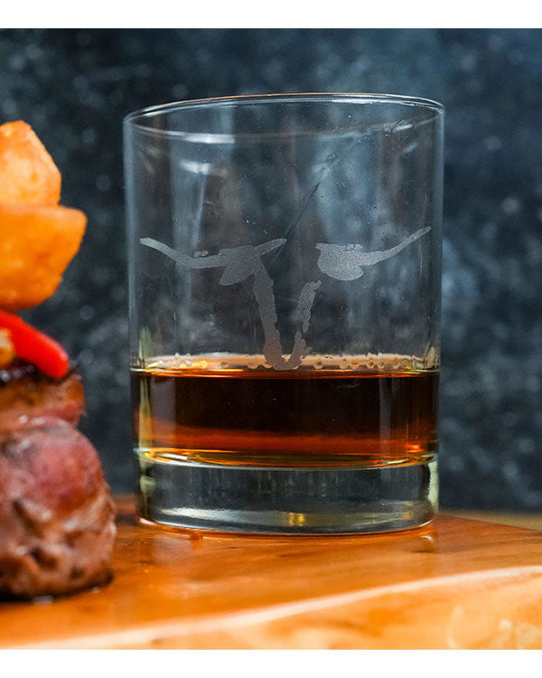 A smooth whiskey glass with amber liquid in it and a longhorn head printed on it. The glass is sitting on a wooden surface in front of a black background.