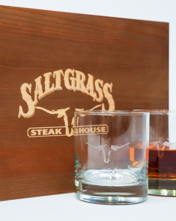 A zoomed in image of a wooden box with "Saltgrass Steak House" printed on it in front of two whiskey glasses with the Saltgrass Longhorn Head in the center, one filled with liquid. The background is plain white.