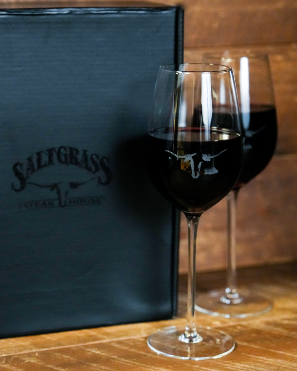 Two wine glasses filled with red wine beside a black box reading "Saltgrass Steak House". The wine glasses both have the Saltgrass Longhorn head printed in the center, as well as the box. The background is a wooden surface and wall.