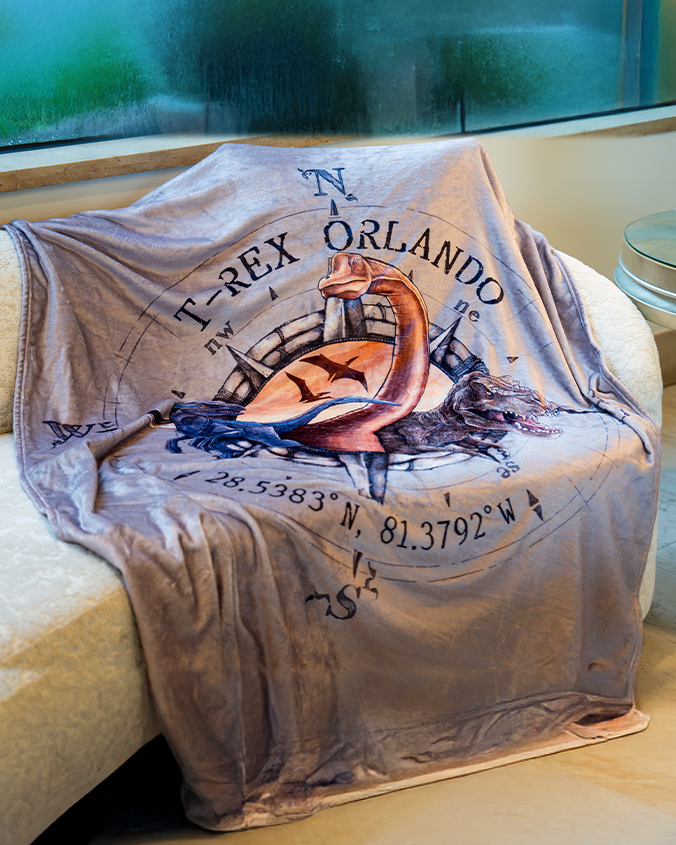 A grey blanket is draped over a beige couch under a foggy window. The blanket has an illustration of three dinosaurs walking out of a compass, with the text "T-REX ORLANDO" written above it, and coordinates around it.