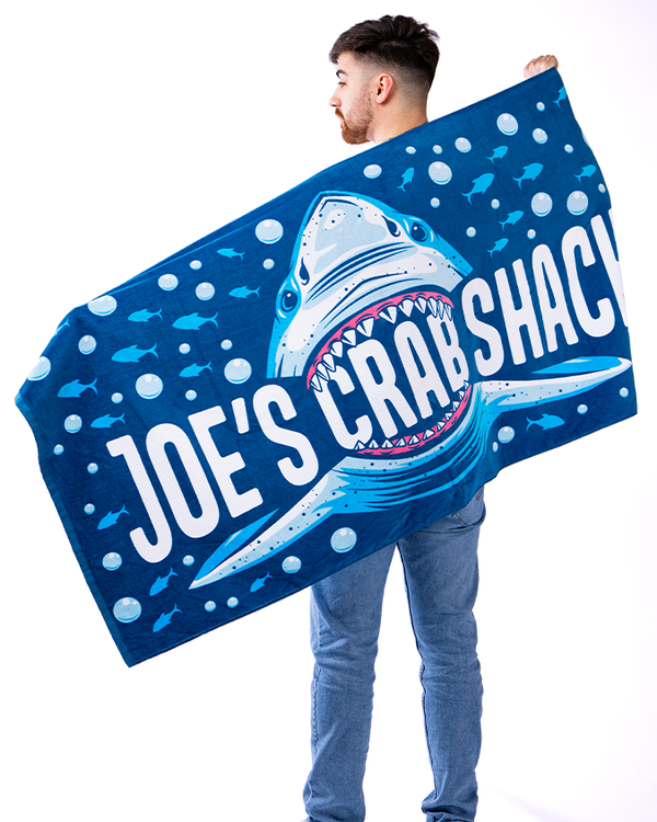 A backside view of a model in blue jeans holding up a blue towel covered with bubbles and fish swimming away from shark with a wide-open mouth, eating the words "JOE'S CRAB SHACK". The background is plain white.