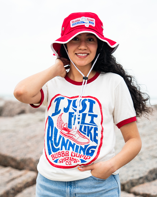 A waist-up view of a dark-haired model in front of a rocky beach with her hand on her neck and one hand tucked into blue jeans, wearing a red bucket hat with white details and adjustable string, including a rectangular Bubba Gump Shrimp Logo. She is also wearing a white baseball t-shirt with red cuff lines, featuring a retro circular design with blue and red wording reading "I Just Felt Like Running" and "Bubba Gump Shrimp Co.", and a shoe with a feather in the middle.
