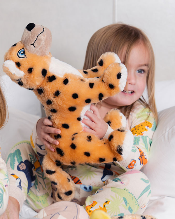 A little girl sitting on a white bed playing with a plush toy jaguar with spots. She is wearing a matching pajama set with leaves and animal patterns.