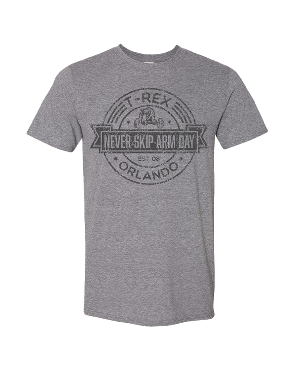 A full view of a gray T-shirt with a circular emblem in the center with the text "T-REX" at the top, "ORLANDO" at the bottom, and "NEVER SKIP ARM DAY, EST 08" in the center in a black ribbon, with a design of a T-rex lifting weights above it. The background is white.