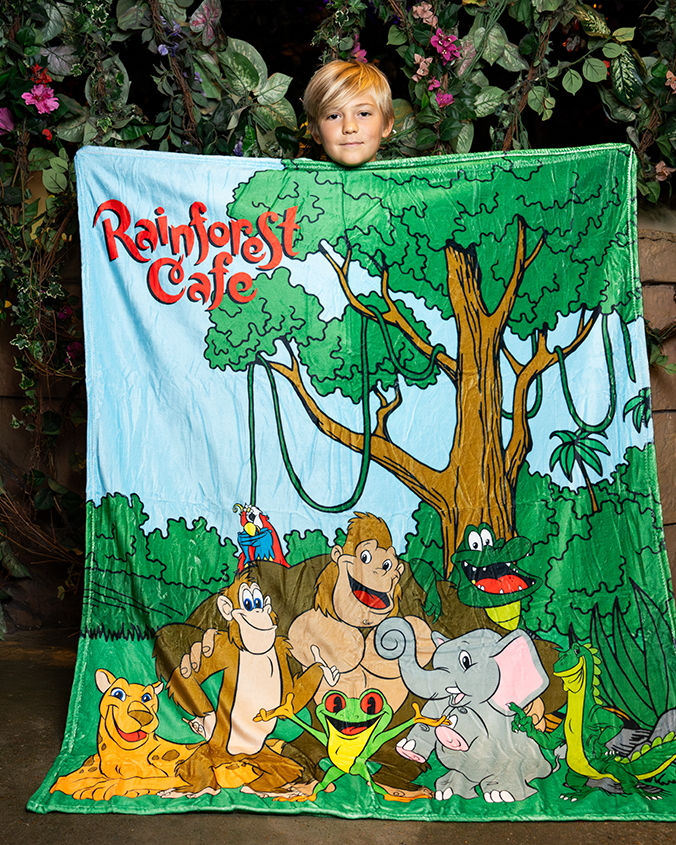 A little boy in a rainforest setting holding up a blanket featuring jungle animals posing in front of a jungle, reading "RAINFOREST CAFE" at the top corner.