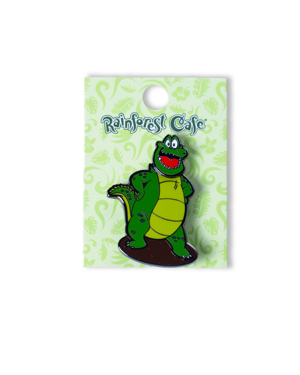 A smiling alligator pin on a brown circle with his hands on his hips, placed on a green card with "RAINFOREST CAFE" on the top. The background is plain white, featuring a rainbow ‘30 WILD YEARS!’ logo featuring CHA! CHA!.