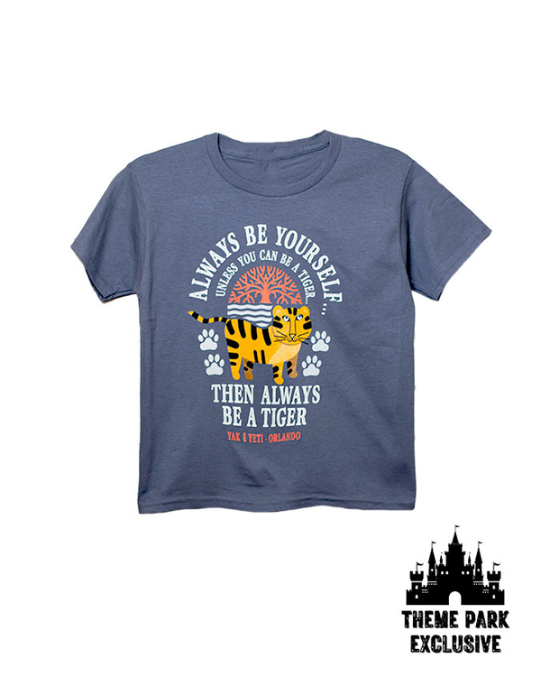 A blue crewneck T-shirt featuring a graphic design of a cartoon tiger surrounded by paw prints with a tree above it, and text that reads 'ALWAYS BE YOURSELF UNLESS YOU CAN BE A TIGER,' and 'THEN ALWAYS BE A TIGER.' At the bottom, the shirt includes the text 'YAK & YETI - ORLANDO'. The background is plain white with a Disney Castle logo at the bottom reading "THEME PARK EXCLUSIVE".