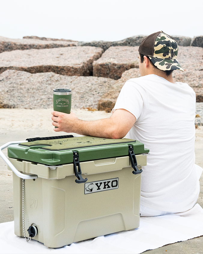 A person sitting on a white towel at the beach, facing away from the camera. The person is holding a green tumbler with "YETI" written on it and wearing a camouflage cap and white t-shirt. Next to the person is a beige cooler with green accents, labeled "YKO,". 