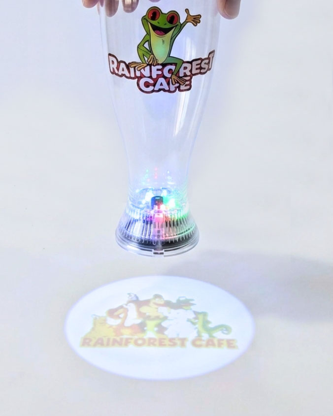A clear plastic party cup with a green frog and 'Rainforest Cafe' logo, held up by hands. The cup has a built-in light-up base that projects a matching circular projection on the floor.