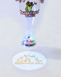 A clear plastic party cup with a green frog and 'Rainforest Cafe' logo, held up by hands. The cup has a built-in light-up base that projects a matching circular projection on the floor.