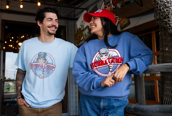 Two casually dressed individuals stand together, wearing branded Bubba Gump merchandise, with a rustic restaurant ambiance in the background.