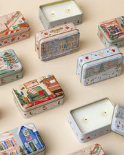 A collection of decorative rectangular tins with various illustrations on their lids, arranged on a beige surface. The tins feature festive designs including buildings, people, and holiday motifs like trees and stars. Some tins are open, showing candle with two wicks.