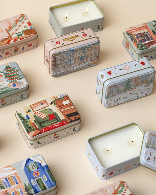 A collection of decorative rectangular tins with various illustrations on their lids, arranged on a beige surface. The tins feature festive designs including buildings, people, and holiday motifs like trees and stars. Some tins are open, showing candle with two wicks.