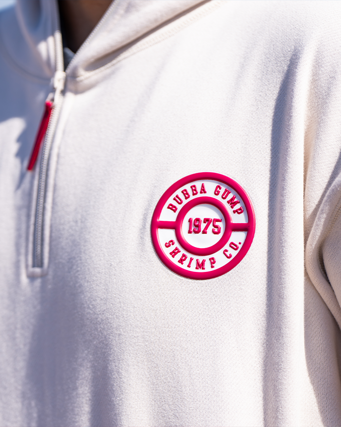 A close-up image of a models' chest wearing a white 3/4 red zippered hoodie with a red circular embroidered emblem on the chest pocket area, reading "BUBBA GUMP SHRIMP CO." and "1975" in the middle. 