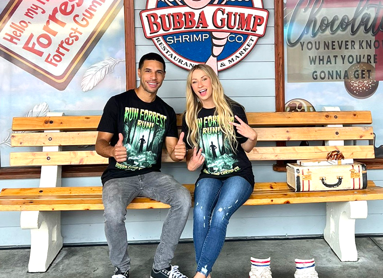  Two individuals are seated on a bench in front of the Bubba Gump Shrimp Co. restaurant. The person on the left is wearing a black t-shirt with green text and the person on the right is wearing a black t-shirt with black text. In front of them is a wooden table with what appears to be a box of chocolates and a sign that reads “Life is like a box of chocolates… you never know what you’re gonna get.” 