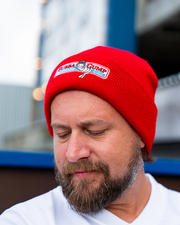 A person wearing a red beanie with an embroidered patch on the front. The patch features a cartoon shrimp character and text that reads "BUBBA GUMP." The background includes an out-of-focus building and lights.