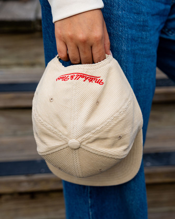 A close-up view of a model's hand holding a beige corduroy baseball cap with red embroidered cursive text on the back reading "Mitchell & Ness". The model is wearing blue jeans and a white shirt, and standing in front of a set of wooden stairs.