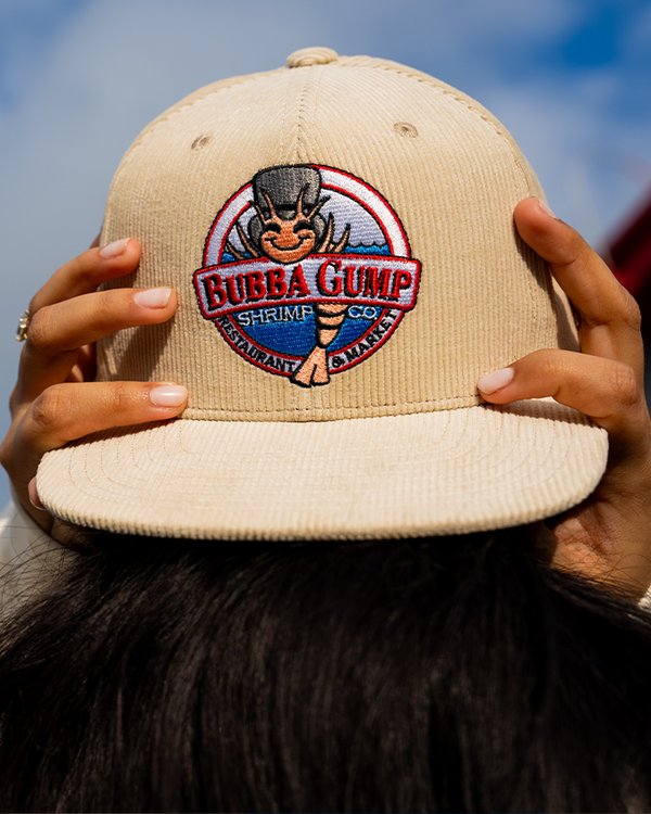 A close-up view of a brunette model standing in front of the sky, wearing and holding onto a beige cap, which has an embroidered logo reading "BUBBA GUMP SHRIMP CO. RESTAURANT & MARKET" with an illustration of a shrimp wearing a hat in front of the ocean. 