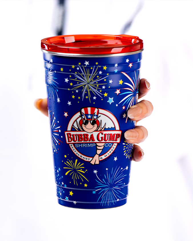 A blue tumbler designed as a solo cup with a red cap, and a body adorned with white stars and fireworks designs. In the center of the cup is a circular “BUBBA GUMP SHRIMP CO.”, featuring a Shrimp in an American flag hat and sunglasses. The background is plain white.