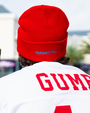 A backside chest-up view of a model in a white jersey reading "GUMP" in red bold text and a red beanie with blue embroidered text on the back reading "MITCHELL & NESS". The background is a blurry building and trees.