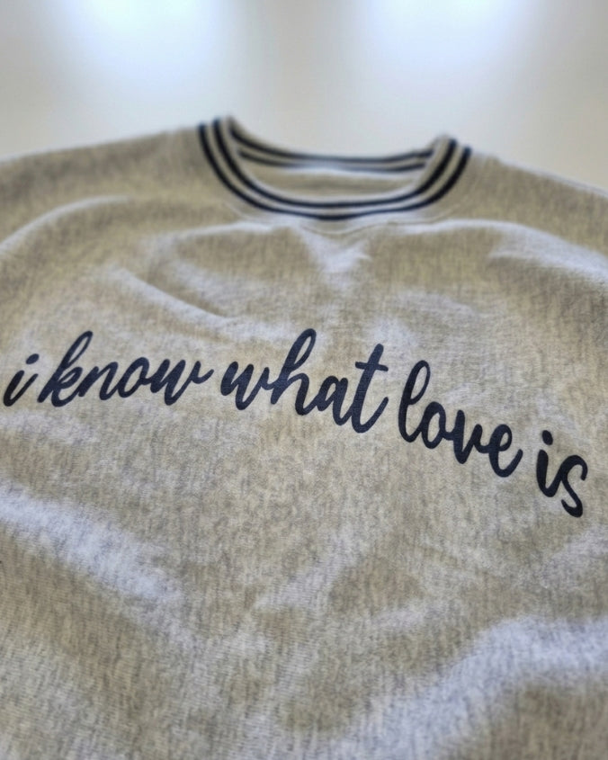A close-up of a light heather gray sweatshirt with navy blue text reading 'i know what love is' across the chest, featuring a striped collar.