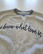 A close-up of a light heather gray sweatshirt with navy blue text reading 'i know what love is' across the chest, featuring a striped collar.