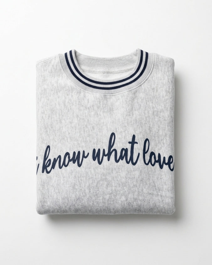 A folded light heather gray sweatshirt with navy blue trim on the crew neck and navy blue script text reading 'I know what love' across the front, shown on a clean white background.