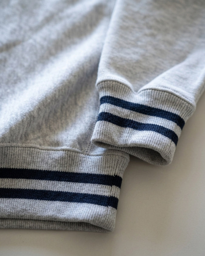 Close-up of a light heather gray sweatshirt with navy blue stripes on the ribbed cuffs, laid flat on a light neutral surface.