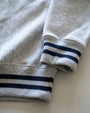 Close-up of a light heather gray sweatshirt with navy blue stripes on the ribbed cuffs, laid flat on a light neutral surface.