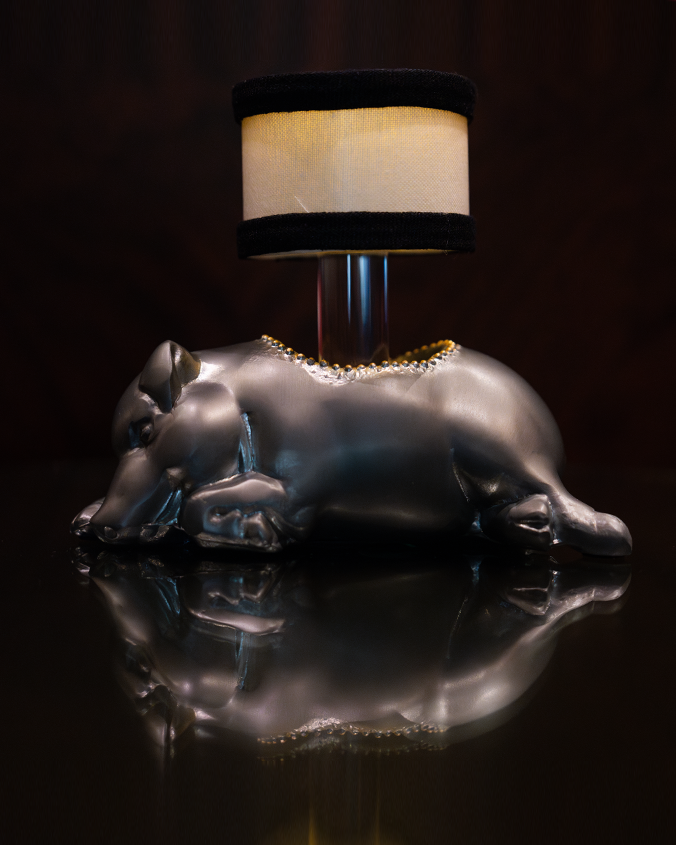 A sideview of a lamp with a silver base shaped like a sleeping pig with a hole in the middle, and a cylindrical lampshade with black trim at the top and bottom coming out of the it. The background is plain black.