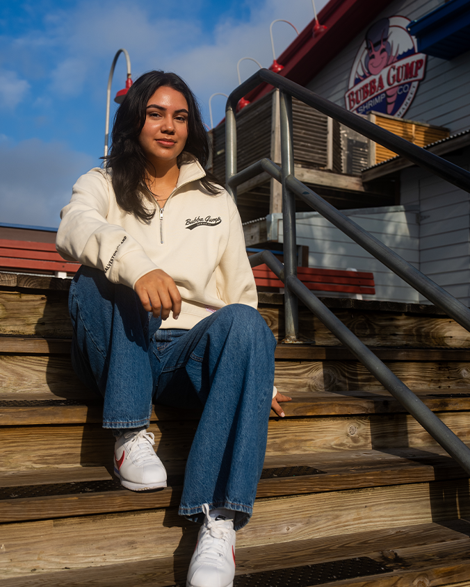 A model sitting on wooden steps behind a Bubba Gump Shrimp Restaurant wearing a cream-colored 3/4 zip sweater with wording on the left chest reading "BUBBA GUMP" and wording on the arm cuff. She is also wearing blue jeans, and white sneakers.