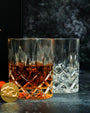 Two crystal whiskey glasses on a dark surface, one filled with amber liquid and the other empty. The glasses feature a diamond-cut pattern that reflects light, creating a sparkling effect. In the background, there is a blurred dark backdrop with light reflecting off its textured surface. 