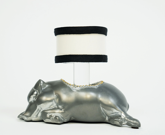 A metallic silver sculpture of a laying pig with a flat surface on its back supporting a cylindrical white lampshade with black bands at the top and bottom. The lampshade is positioned where the pig's back would naturally rise, turning the sculpture into a functional lamp.