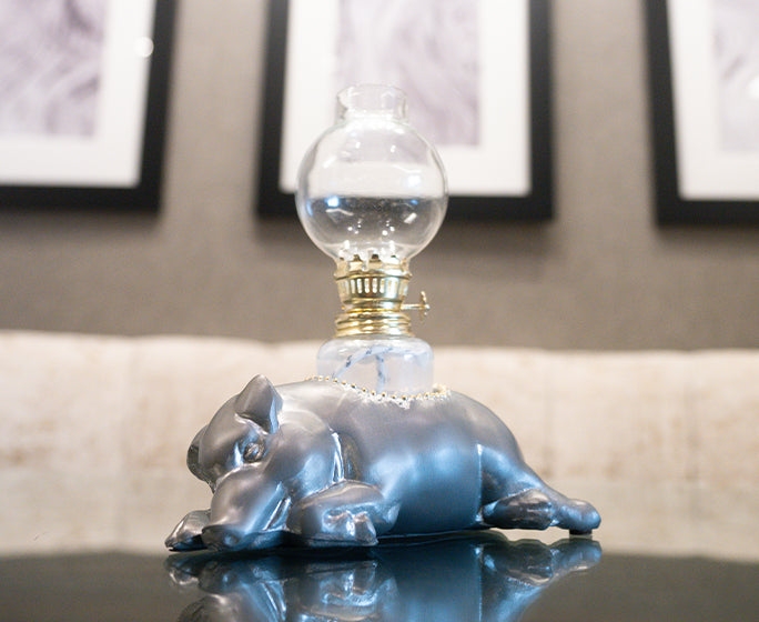 A unique lamp with a base shaped like a lying down pig, in a glossy blue finish. The lamp has a clear glass shade on top. In the background, there are framed artworks hanging on a wall with a patterned wallpaper. The lamp is placed on a reflective surface, possibly a table, and the setting suggests an interior home decor environment.